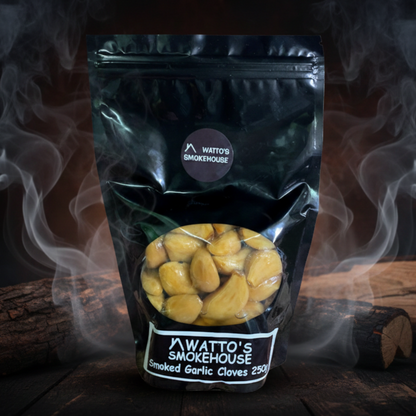 Package of smoked garlic cloves with 'Watto's Smokehouse' branding, surrounded by smoke and wooden logs on a dark background.